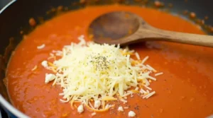 Grated cheese and ricotta being added to a rich tomato sauce in a skillet, with a wooden spoon resting on the side.