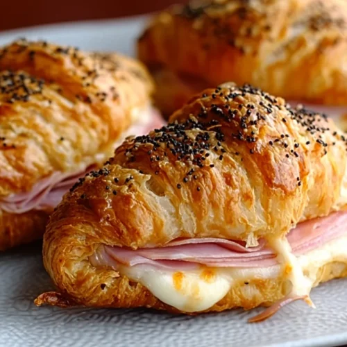 Golden brown savory baked ham and cheese croissants on a white plate, filled with melted cheese and sliced ham, topped with poppy seeds.