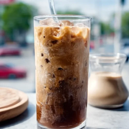 Tall glass of creamy iced coffee with swirling milk and ice cubes on a light table.
