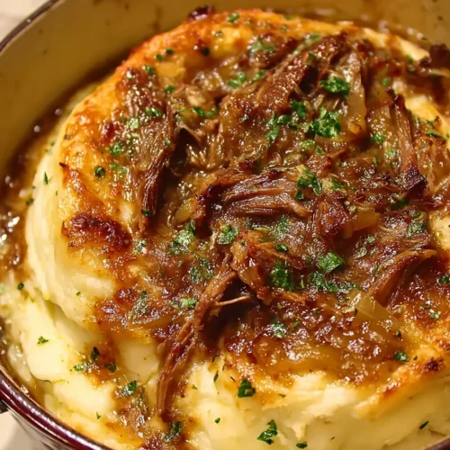 French onion pot roast served over creamy mashed potatoes in a brown bowl, topped with caramelized onions, fresh herbs, and a sprig of rosemary, with slices of rustic bread in the background.