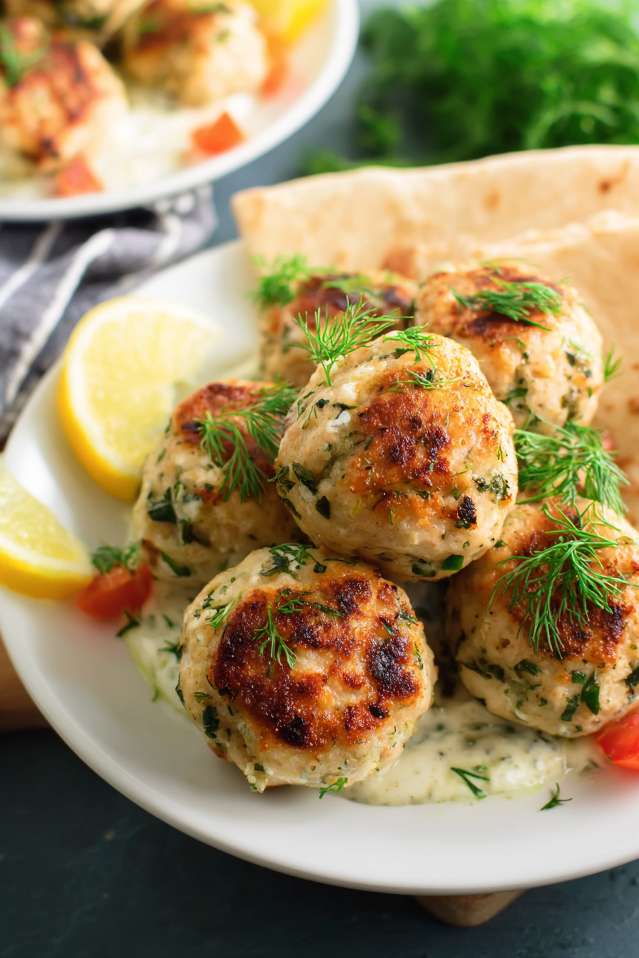 Mediterranean chicken meatballs garnished with fresh dill served on a white plate with tzatziki sauce, lemon wedges, cherry tomatoes, and warm pita bread