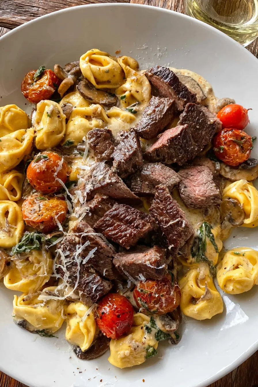 Garlic steak bites served over creamy tortellini with mushrooms and roasted cherry tomatoes on a white plate