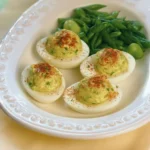 Deviled eggs topped with green herb filling and paprika on a white plate with green beans in the background